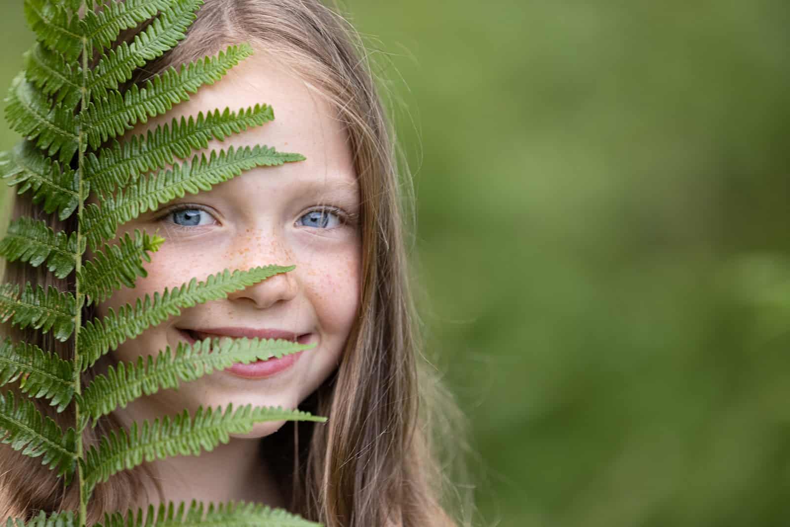 Child smiles at the camera, peeking out from behind a fern leaf during a photoshoot in Hemel Hemstead